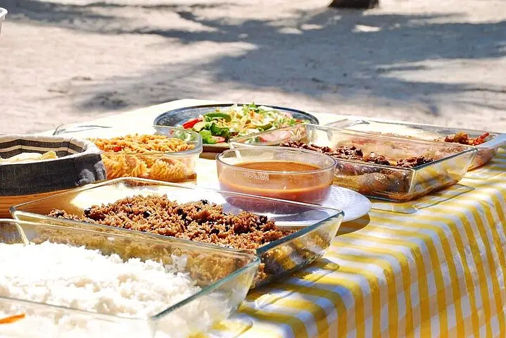 A picnic table with a yellow checkered tablecloth is set outdoors, displaying dishes of rice, pasta, salad, stew, and meat. Sunlight casts shadows across the table, creating a bright atmosphere perfect for enjoying between the best tours Punta Cana offers.