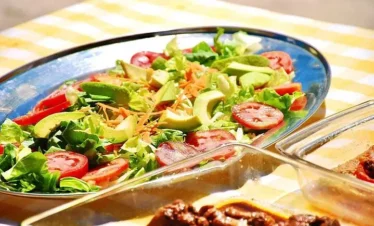 A colorful salad with sliced tomatoes, avocado, lettuce, and shredded carrots sits on a shiny oval platter. In the foreground, a glass dish with a brown cooked dish is slightly blurred—a scene perfect after enjoying the best tours in Punta Cana.