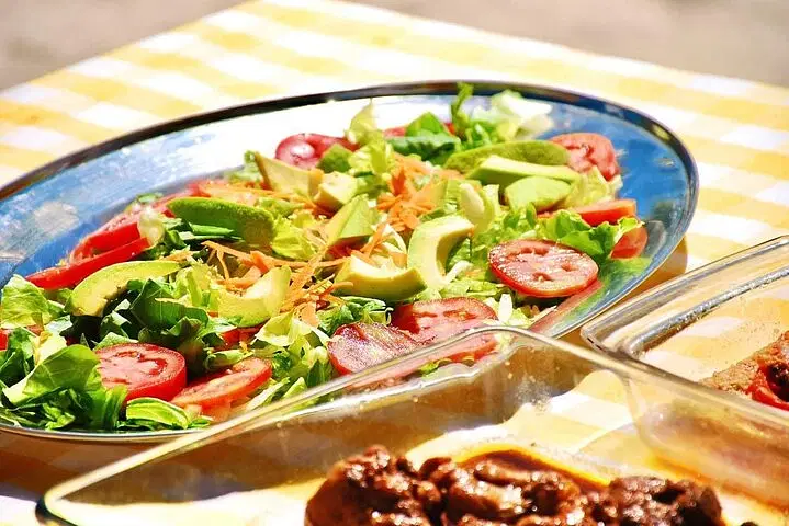 A colorful salad with sliced tomatoes, avocado, lettuce, and shredded carrots sits on a shiny oval platter. In the foreground, a glass dish with a brown cooked dish is slightly blurred—a scene perfect after enjoying the best tours in Punta Cana.