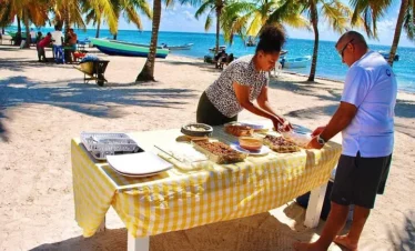 Two people serve food from a yellow-checked table on a sandy beach, under palm trees. Nearby boats rest by the turquoise sea. Plates and trays are neatly arranged—just one highlight you might find on tours Punta Cana has to offer.