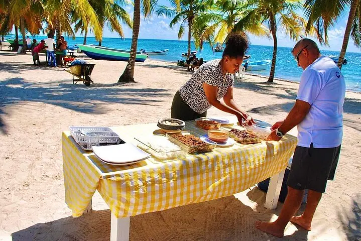 Two people serve food from a yellow-checked table on a sandy beach, under palm trees. Nearby boats rest by the turquoise sea. Plates and trays are neatly arranged—just one highlight you might find on tours Punta Cana has to offer.