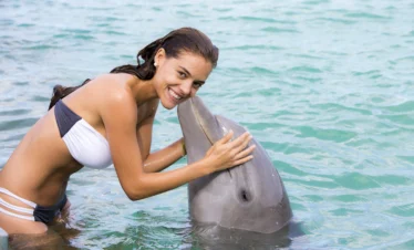 A smiling woman in a white bikini hugs and interacts with a dolphin in clear, turquoise water during one of the unforgettable Punta Cana excursions. She leans forward, holding the dolphin as it affectionately touches her face above the surface.