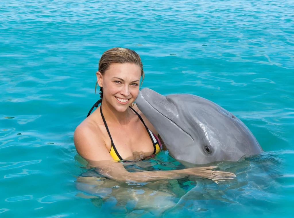 A smiling woman in a bikini top stands in clear blue water during one of the popular Punta Cana excursions, hugging a dolphin. The dolphin presses its snout affectionately to her cheek as they enjoy the sparkling turquoise sea together.