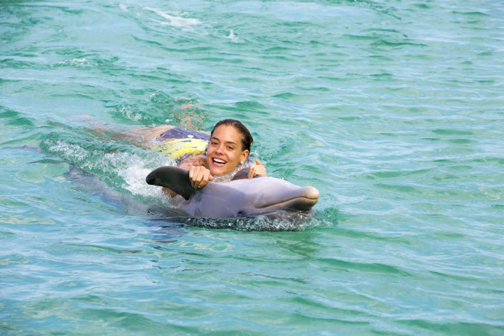 A smiling woman in a yellow swimsuit holds onto a dolphin's fin, being pulled through clear, turquoise water during her Punta Cana trip. She looks joyful and relaxed as the dolphin swims ahead, creating playful splashes around them.