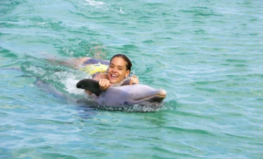 A smiling woman in a yellow swimsuit holds onto a dolphin's fin, being pulled through clear, turquoise water during her Punta Cana trip. She looks joyful and relaxed as the dolphin swims ahead, creating playful splashes around them.