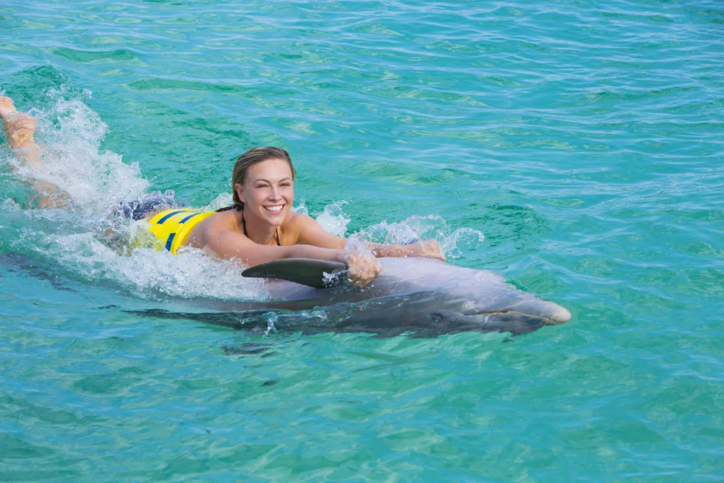 A smiling woman in a yellow life jacket holds onto a dolphin’s fin, riding it through clear turquoise water. The bright, sunny atmosphere highlights the joy of this unforgettable Punta Cana excursion.