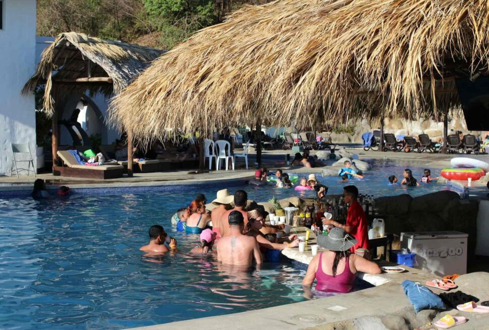 People relax in a pool with a swim-up bar under a thatched roof in Punta Cana. Some sit at the bar, others swim or float on inflatable rings. White plastic chairs and umbrellas dot this sunny, lively scene of budget-friendly thrills for adults and children alike.