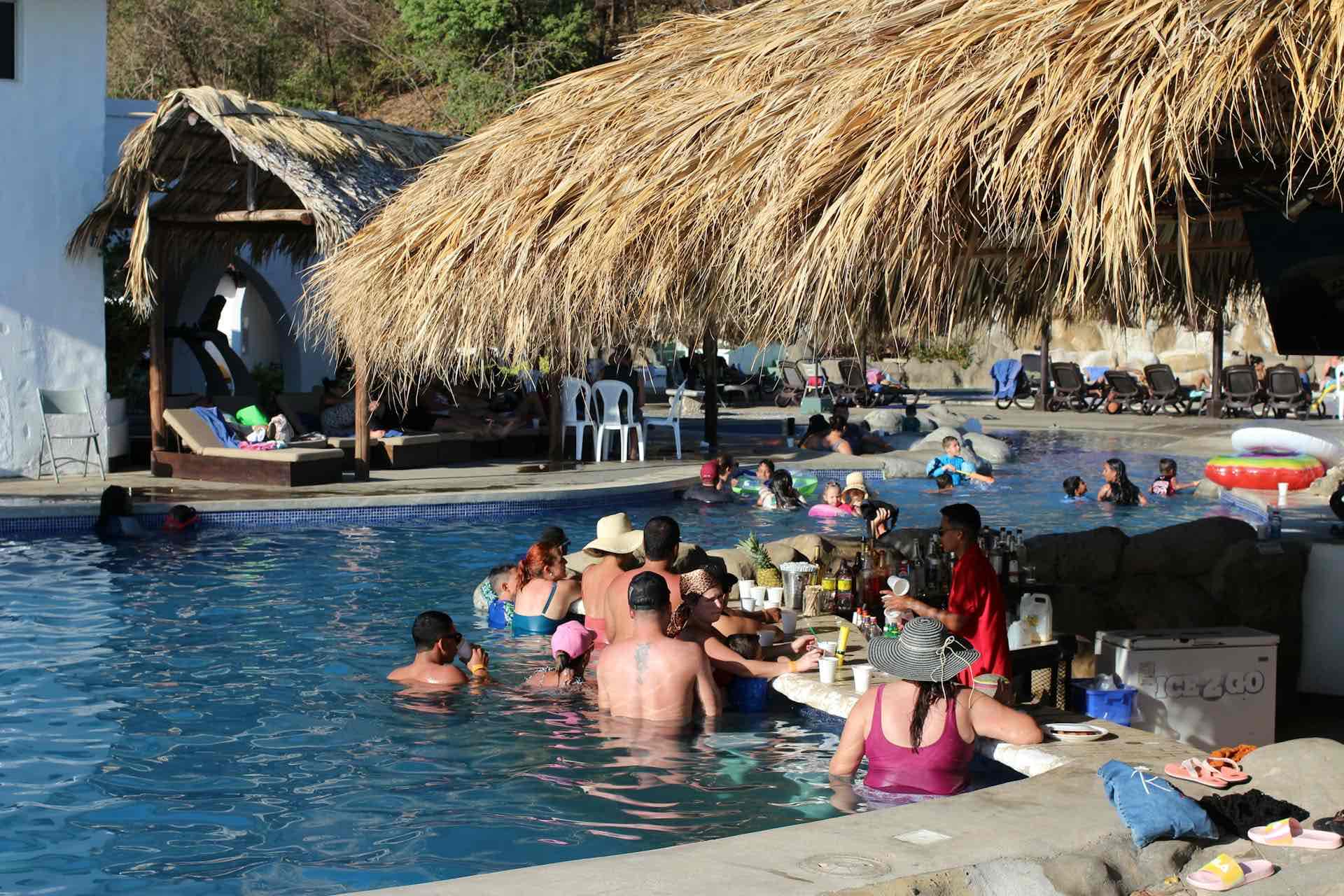People relax in a pool with a swim-up bar under a thatched roof in Punta Cana. Some sit at the bar, others swim or float on inflatable rings. White plastic chairs and umbrellas dot this sunny, lively scene of budget-friendly thrills for adults and children alike.
