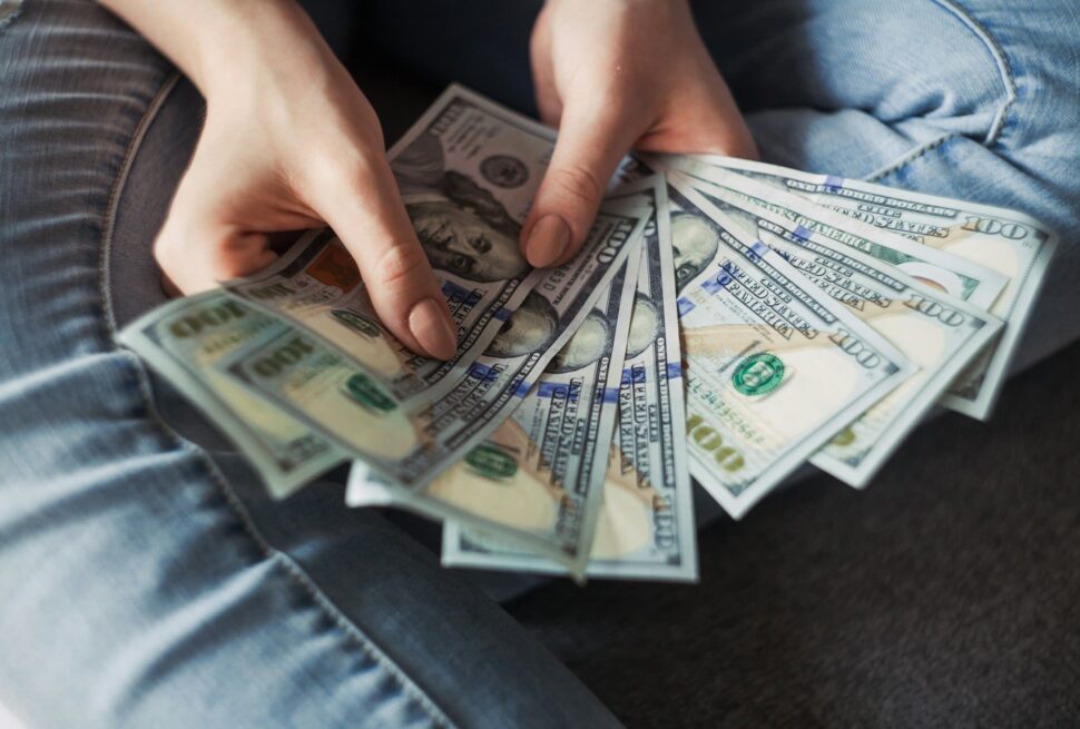 A person in blue jeans sits cross-legged, holding a fan of US $100 bills in both hands, showcasing their fronts—suggesting financial abundance, travel spending, or mindful handling of a vacation budget. The softly blurred background keeps the focus on the money.