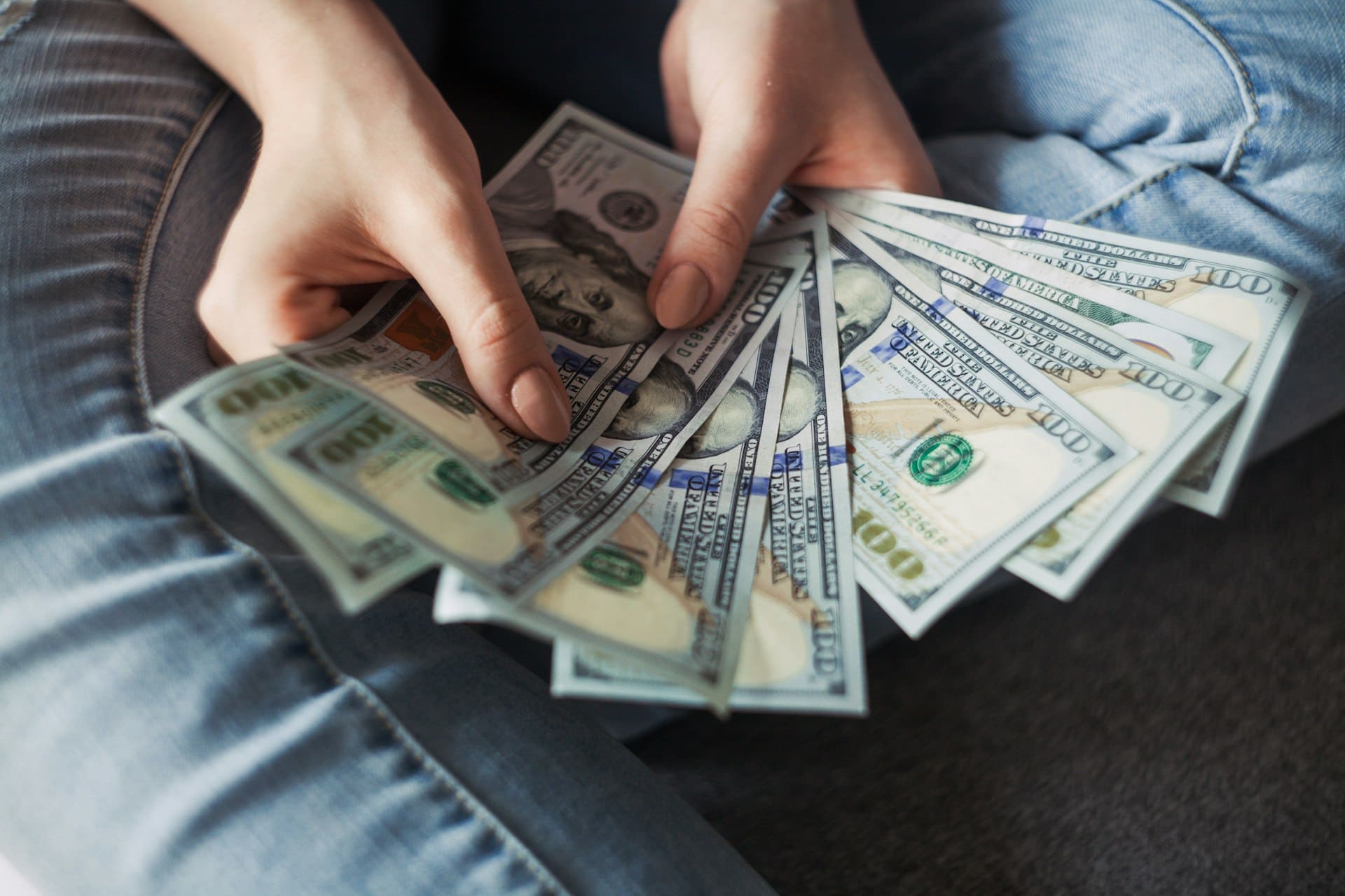 A person in blue jeans sits cross-legged, holding a fan of US $100 bills in both hands, showcasing their fronts—suggesting financial abundance, travel spending, or mindful handling of a vacation budget. The softly blurred background keeps the focus on the money.