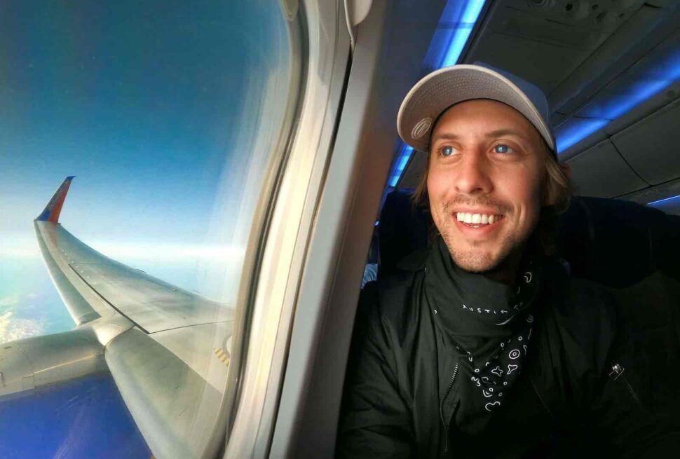 A smiling man traveling alone, wearing a light cap, black jacket, and bandana, sits by an airplane window. Sunlight illuminates his face as he enjoys the pros and cons of solo travel. Outside, a wing cuts through a sky dotted with scattered clouds.