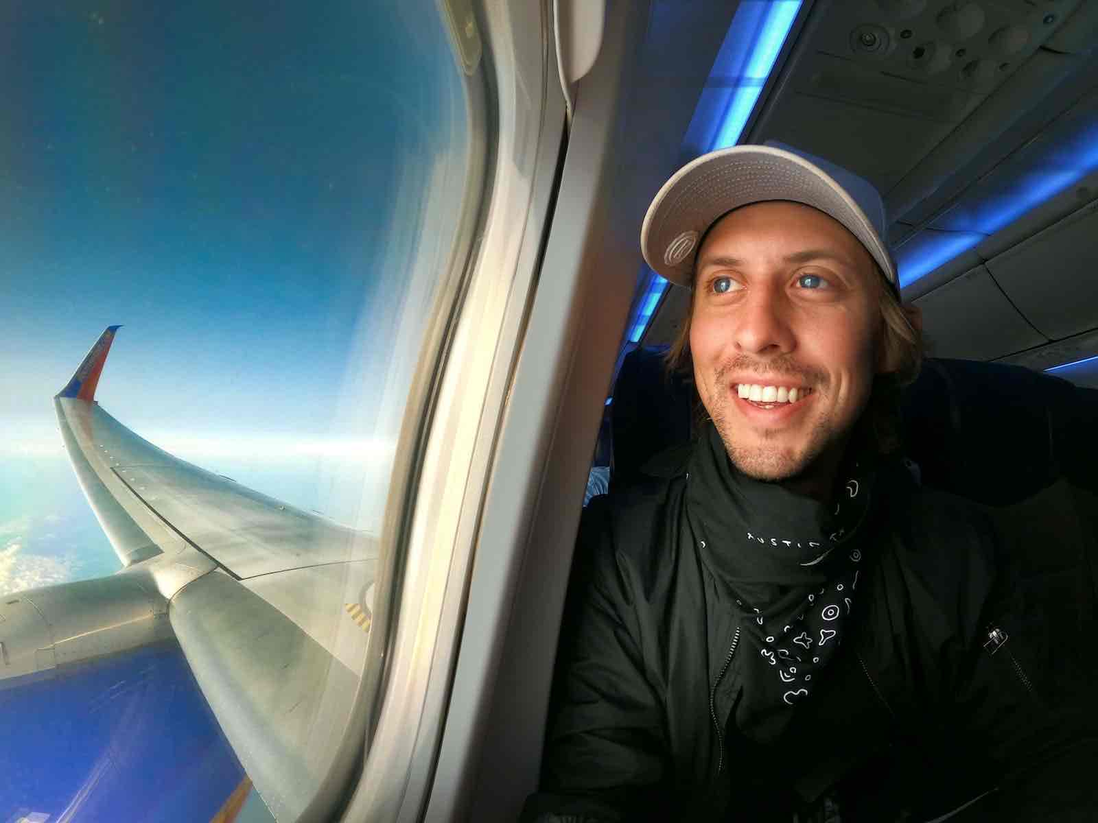 A smiling man traveling alone, wearing a light cap, black jacket, and bandana, sits by an airplane window. Sunlight illuminates his face as he enjoys the pros and cons of solo travel. Outside, a wing cuts through a sky dotted with scattered clouds.