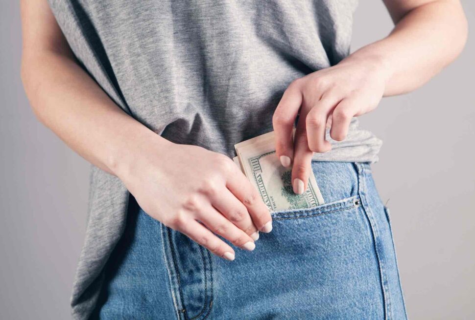 A person wearing a grey t-shirt and blue jeans places a folded stack of U.S. hundred dollar bills into their front jeans pocket, perhaps saving up for all-inclusive holidays. Only the hands, torso, and upper jeans are visible against a plain background.