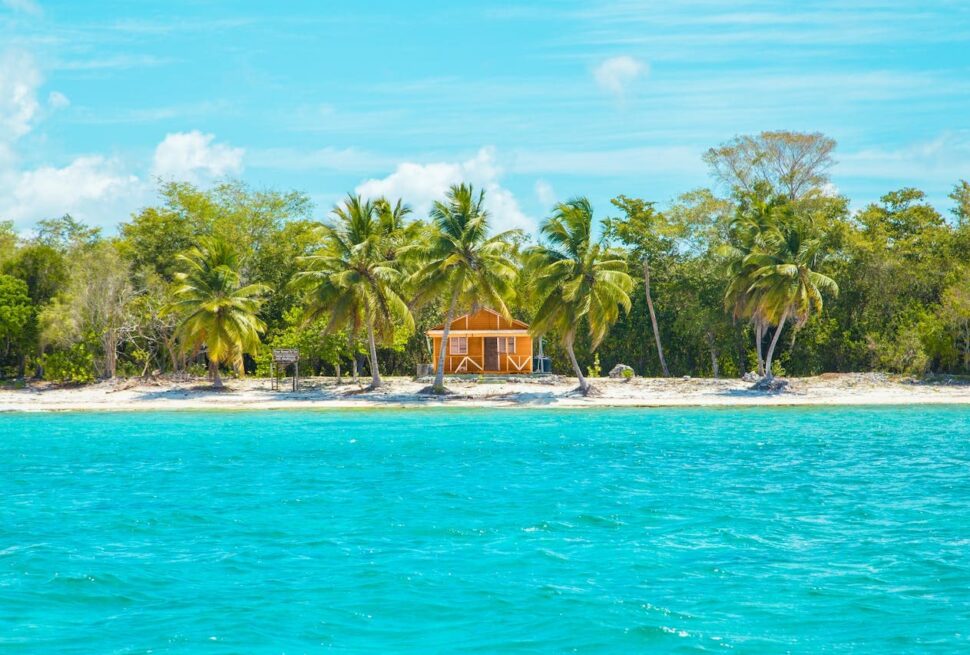A small yellow beach house with a porch is surrounded by palm trees and dense greenery on a sandy Punta Cana shoreline. Clear turquoise water shimmers in the foreground under a bright, sunny sky, offering budget-friendly thrills.
