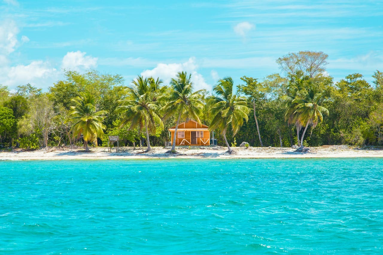 A small yellow beach house with a porch is surrounded by palm trees and dense greenery on a sandy Punta Cana shoreline. Clear turquoise water shimmers in the foreground under a bright, sunny sky, offering budget-friendly thrills.