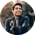 A young man with dark hair smiles while standing outdoors during tours Punta Cana. He wears a dark jacket over a shirt and has his arms crossed. The softly blurred background hints at natural Punta Cana scenery in brown and green tones, with a circular crop.