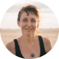 A woman with light skin and brown hair smiles gently at the camera, wearing a black tank top and turquoise pendant. Behind her, a sandy beach and cloudy sky evoke the serene beauty often discovered on Punta Cana tours.