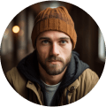 A young man with a brown knit beanie and short beard looks directly at the camera, ready for adventure. He wears a tan jacket over a dark hoodie and gray shirt—looking like someone planning the best tours or seeking smooth Punta Cana transfers.