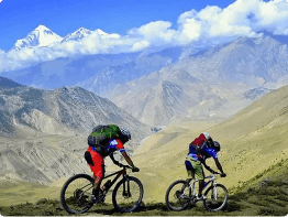Two cyclists with backpacks ride mountain bikes uphill on a rugged trail. Dramatic, steep hills and snow-capped peaks set the scene for adventure—like the best tours, it captures the thrill and challenge of exploring remote, high-altitude landscapes.
