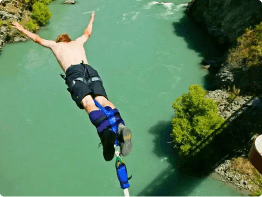 A person with short hair, shirtless and wearing shorts, is mid-air in a bungee jump over a turquoise river—arms outstretched and harnessed legs—capturing the thrill of the best tours as rocky cliffs and greenery stretch below.