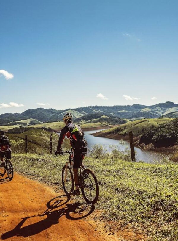 Two cyclists ride on a winding dirt road alongside a river, surrounded by green hills under a clear blue sky. The lush grass borders the route—just like the scenic trails you’ll discover with Punta Cana tours or the best tours in the region.