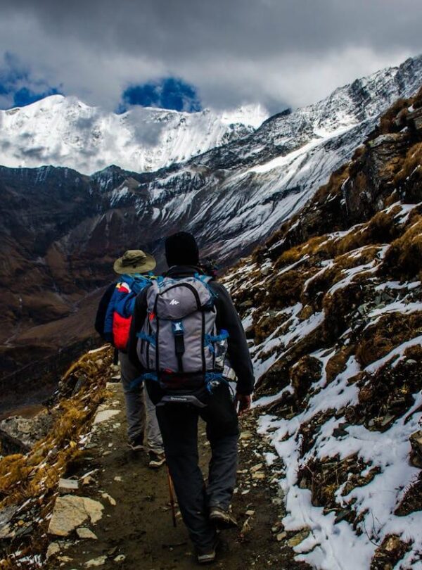 Two hikers with backpacks walk a narrow, rocky mountain trail past snowy slopes and majestic peaks under dramatic clouds, capturing the spirit of adventure found on the best tours—far from sunny Punta Cana tours or convenient Punta Cana transfers.