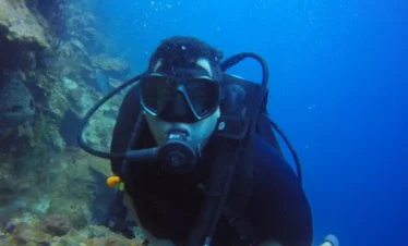A scuba diver wearing black diving gear, a mask, and yellow fins swims underwater near a rocky coral reef on a Punta Cana trip. Bubbles rise from the diver's regulator as they look toward the camera, surrounded by clear, blue water.