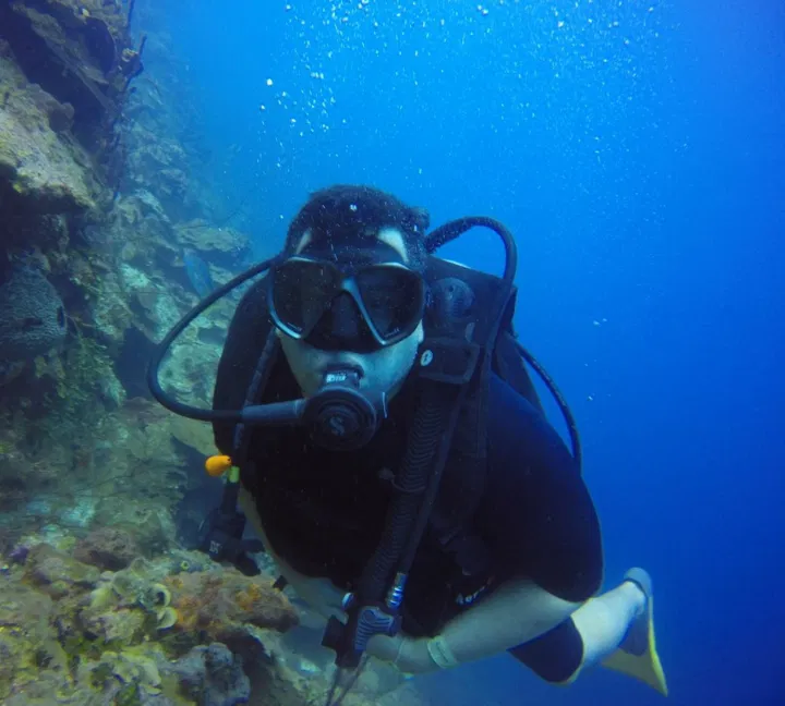 A scuba diver wearing black diving gear, a mask, and yellow fins swims underwater near a rocky coral reef on a Punta Cana trip. Bubbles rise from the diver's regulator as they look toward the camera, surrounded by clear, blue water.
