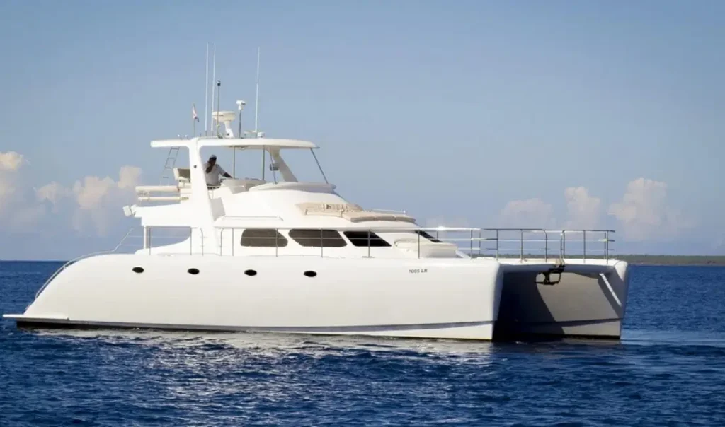 A large, white catamaran yacht floats on calm blue water under a clear sky—perfect for Punta Cana excursions. With two hulls, multiple windows, and a spacious upper deck, it cruises toward distant land with fluffy clouds on the horizon.