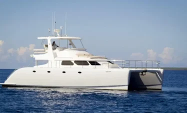 A large, white catamaran yacht floats on calm blue water under a clear sky—perfect for Punta Cana excursions. With two hulls, multiple windows, and a spacious upper deck, it cruises toward distant land with fluffy clouds on the horizon.