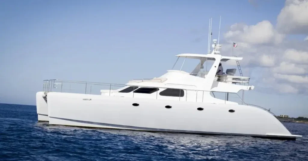 A sleek white catamaran yacht floats on calm blue ocean water under a partly cloudy sky. Perfect for Punta Cana excursions, the boat has multiple windows, a covered upper deck, and railings. Land is barely visible in the distance on the right side.