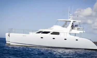 A sleek white catamaran yacht floats on calm blue ocean water under a partly cloudy sky. Perfect for Punta Cana excursions, the boat has multiple windows, a covered upper deck, and railings. Land is barely visible in the distance on the right side.