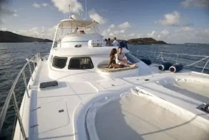 A large white yacht is cruising on calm water under a partly cloudy sky. Two women relax on the front deck, chatting, as others enjoy the upper deck. Small islands dot the background—an ideal setting for exclusive Punta Cana tours and island leisure.