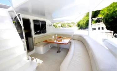 A bright, modern yacht deck features an L-shaped cream leather sofa, a wooden table set for a meal, and a staircase leading upward—perfect for relaxing after Punta Cana excursions, with large windows showcasing lush greenery and another boat on calm water.