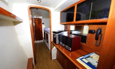 A narrow room with wood paneling, cabinets, and shelving. A desk holds a printer, phone, folders, and papers—perfect for planning Punta Cana tours. A doorway reveals striped flooring and a bed beyond. Warm lighting creates a cozy atmosphere.