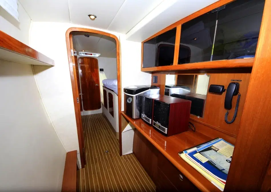 A narrow room with wood paneling, cabinets, and shelving. A desk holds a printer, phone, folders, and papers—perfect for planning Punta Cana tours. A doorway reveals striped flooring and a bed beyond. Warm lighting creates a cozy atmosphere.
