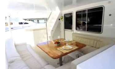 A yacht's outdoor seating area with cream-colored cushioned benches surrounds a wooden dining table, beautifully set for a meal. Large windows and bright natural light create an open, airy feel—perfect for relaxing during your Punta Cana excursions.
