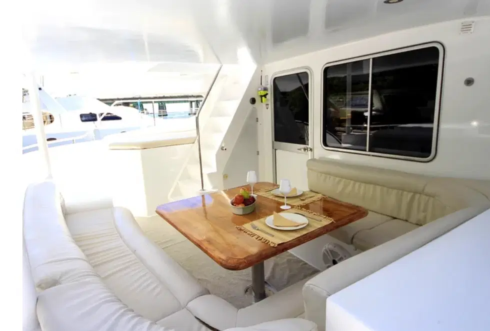 A yacht's outdoor seating area with cream-colored cushioned benches surrounds a wooden dining table, beautifully set for a meal. Large windows and bright natural light create an open, airy feel—perfect for relaxing during your Punta Cana excursions.