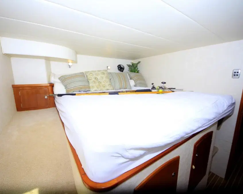 A cozy, compact bedroom features a neatly made bed with white bedding and patterned pillows. Fishing rods rest on the bed, evoking adventure—perfect for relaxing after tours in Punta Cana. Wooden cabinets and beige carpeting suggest the room is inside a boat or RV.