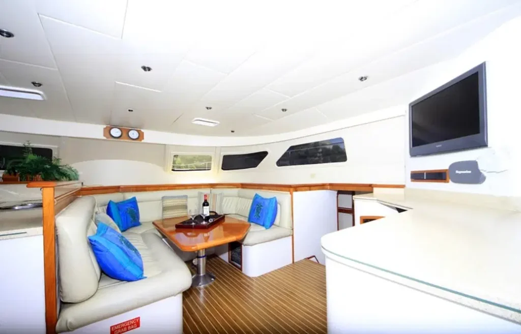 The interior of a yacht, perfect for Punta Cana tours, features a curved cream leather sofa with blue pillows around a wooden table set with wine and glasses. Large windows, a mounted TV, and wood-accented counters create a modern, inviting lounge space.