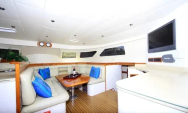 The interior of a yacht, perfect for Punta Cana tours, features a curved cream leather sofa with blue pillows around a wooden table set with wine and glasses. Large windows, a mounted TV, and wood-accented counters create a modern, inviting lounge space.