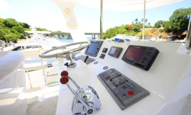 Close-up of a yacht’s helm with a shiny stainless steel steering wheel, throttle levers, various navigation screens, and switches on a white console. The background hints at scenic waters perfect for Punta Cana tours and unforgettable excursions.