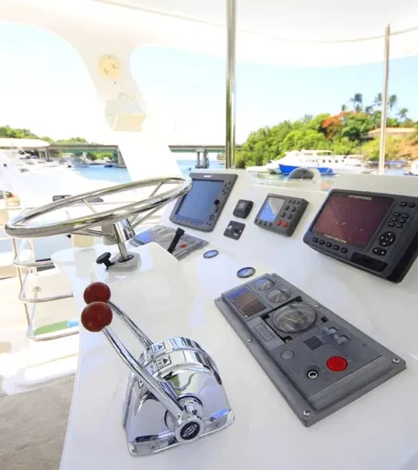 Close-up of a yacht’s helm with a shiny stainless steel steering wheel, throttle levers, various navigation screens, and switches on a white console. The background hints at scenic waters perfect for Punta Cana tours and unforgettable excursions.