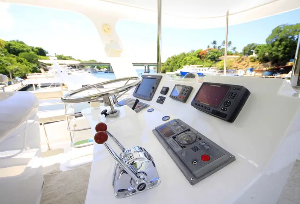 Close-up of a yacht’s helm with a shiny stainless steel steering wheel, throttle levers, various navigation screens, and switches on a white console. The background hints at scenic waters perfect for Punta Cana tours and unforgettable excursions.