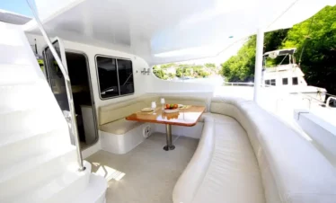 A bright yacht deck features a curved white leather sofa, a wooden table set with plates and fruit, and stairs leading up. Large windows reveal lush greenery and another boat—perfect for luxury tours in Punta Cana with stunning views.