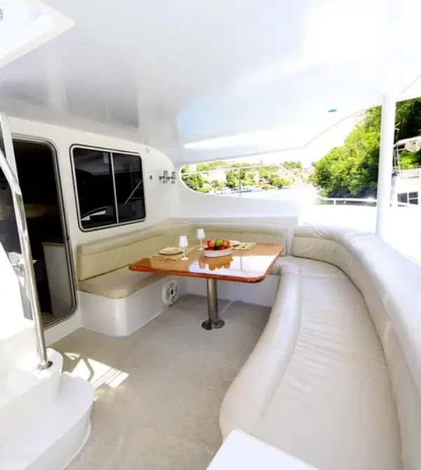 A bright yacht deck features a curved white leather sofa, a wooden table set with plates and fruit, and stairs leading up. Large windows reveal lush greenery and another boat—perfect for luxury tours in Punta Cana with stunning views.