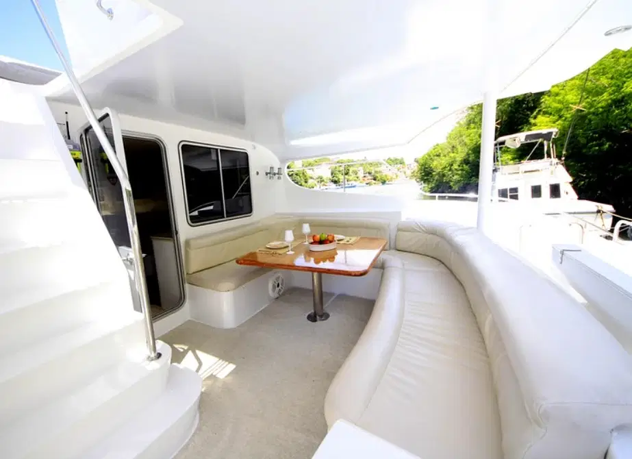 A bright yacht deck features a curved white leather sofa, a wooden table set with plates and fruit, and stairs leading up. Large windows reveal lush greenery and another boat—perfect for luxury tours in Punta Cana with stunning views.