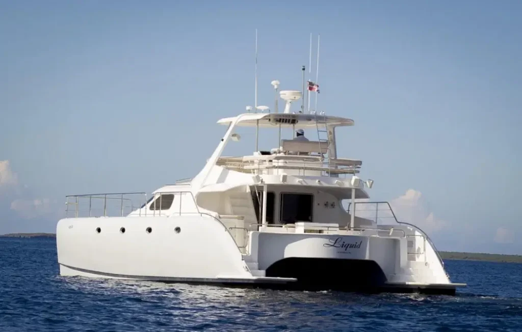 A large white catamaran yacht named "Liquid" floats on calm blue water under a clear sky, ready for unforgettable punta cana tours. The yacht features multiple decks, antennas, railings, wide hulls, and an open aft deck with a small flag on top.