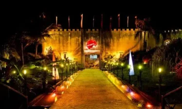 A brightly lit pathway leads to a fortress-like building at night in Punta Cana. Warm yellow lights illuminate the path, with colorful spotlights and triangular light installations. Palms and greenery surround this vibrant scene, perfect for Punta Cana nightlife.