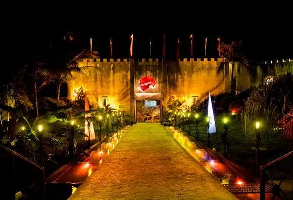A brightly lit pathway leads to a fortress-like building at night in Punta Cana. Warm yellow lights illuminate the path, with colorful spotlights and triangular light installations. Palms and greenery surround this vibrant scene, perfect for Punta Cana nightlife.
